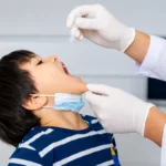 A healthcare professional wearing gloved hands administering oral drops to a young boy with a lowered face mask during a medical procedure.