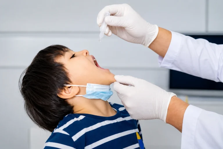 Blog 3 A healthcare professional wearing gloved hands administering oral drops to a young boy with a lowered face mask during a medical procedure.
