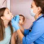 A female doctor wearing a face mask and gloves using a nasal swab to examine a young girl during a clinical consultation.