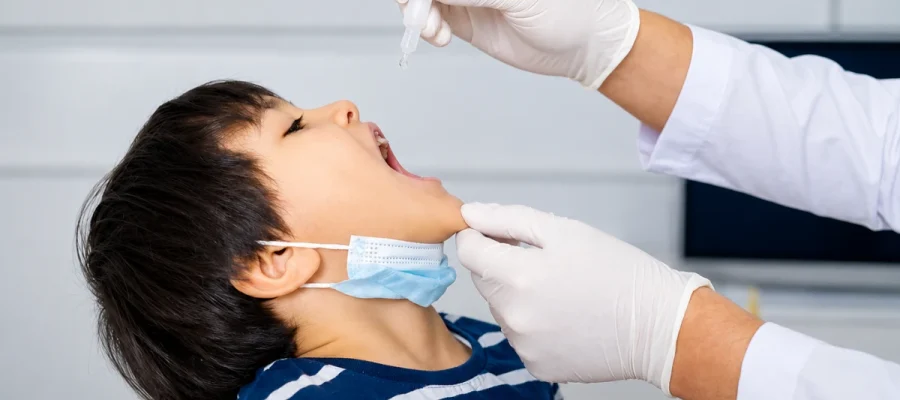 A healthcare professional wearing gloved hands administering oral drops to a young boy with a lowered face mask during a medical procedure.