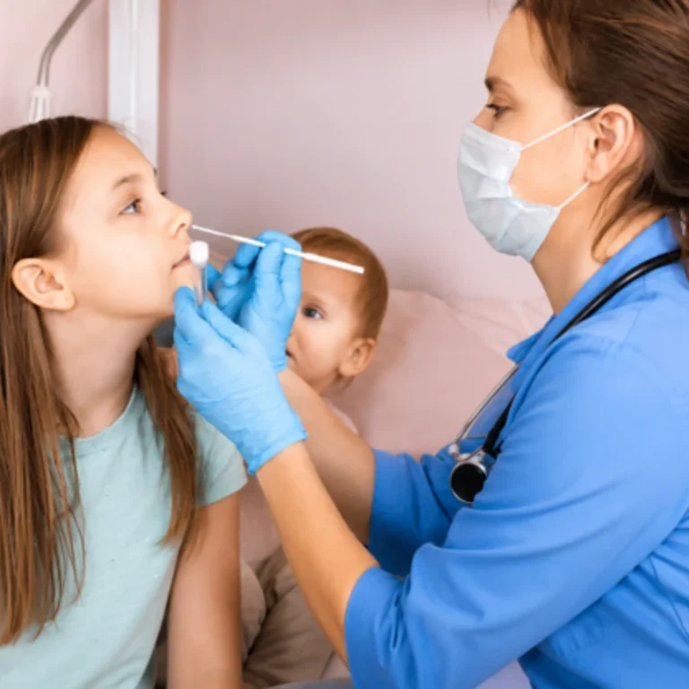A female doctor wearing a face mask and gloves using a nasal swab to examine a young girl during a clinical consultation.